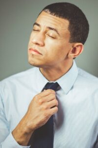 A man indoors adjusting his necktie with a thoughtful expression, wearing formal attire.