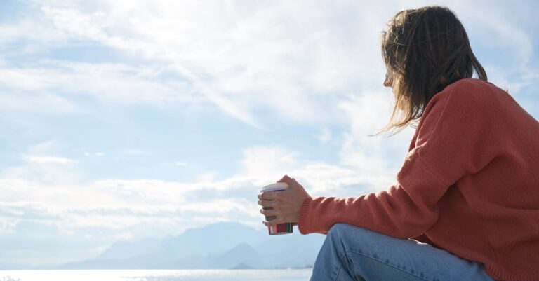 A woman in casual attire enjoys a hot drink while gazing at the sea, epitomizing relaxation.